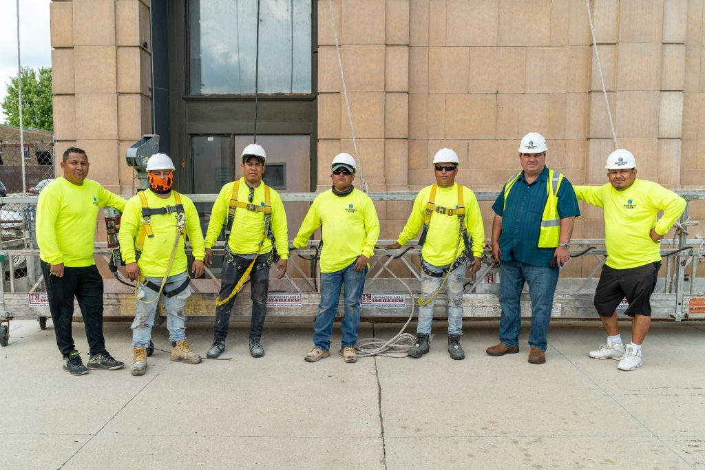 About Us North Shore Masonry crew in hi-vis shirts and hard hats lined up in front of a suspended scaffold platform