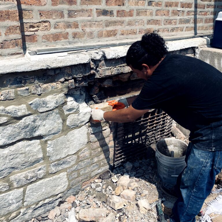About Us Mason in work gloves repairing a stone foundation wall beneath a brick facade